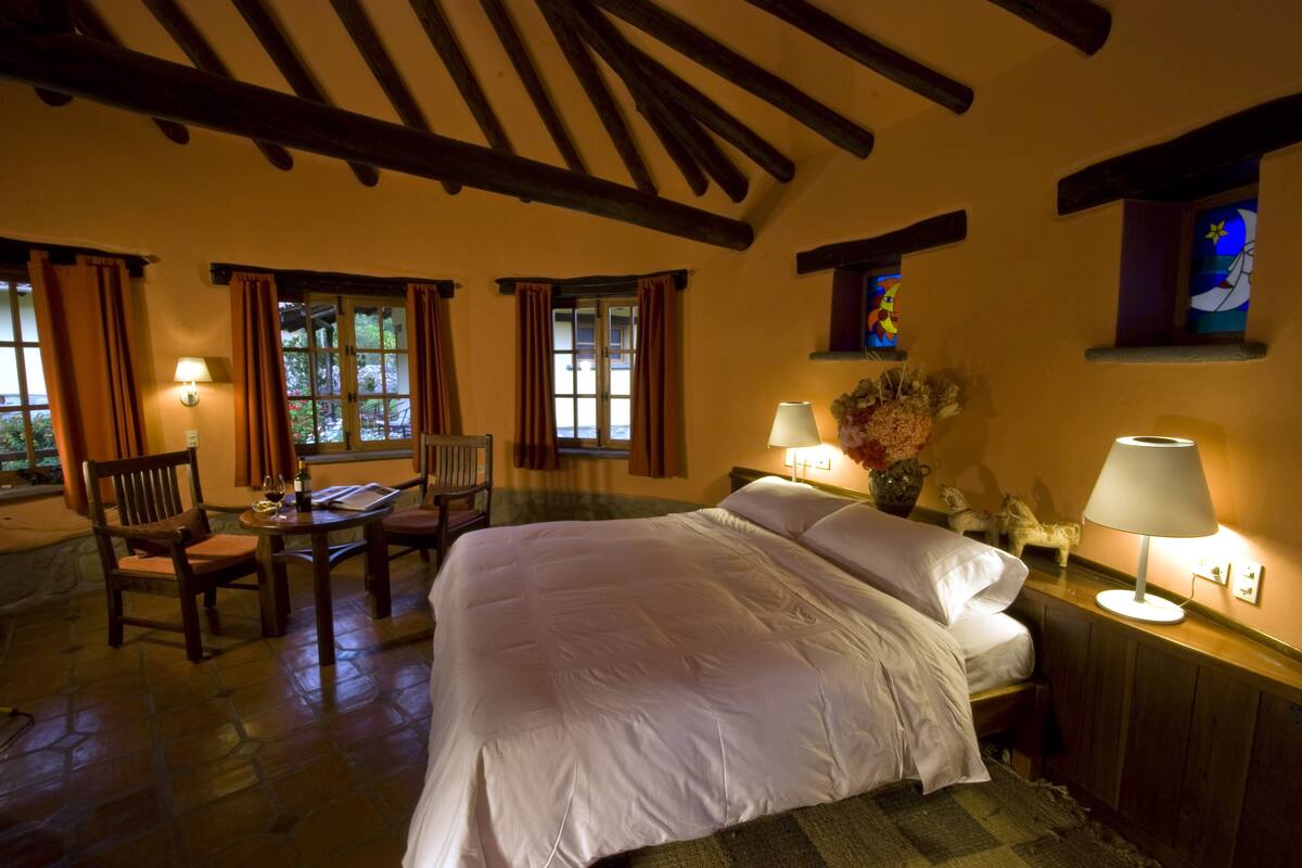 A bedroom with a large bed, two bedside tables with lamps, a chair, and a small table with a chair. There are windows with curtains, a stone floor, and a wooden ceiling beam.