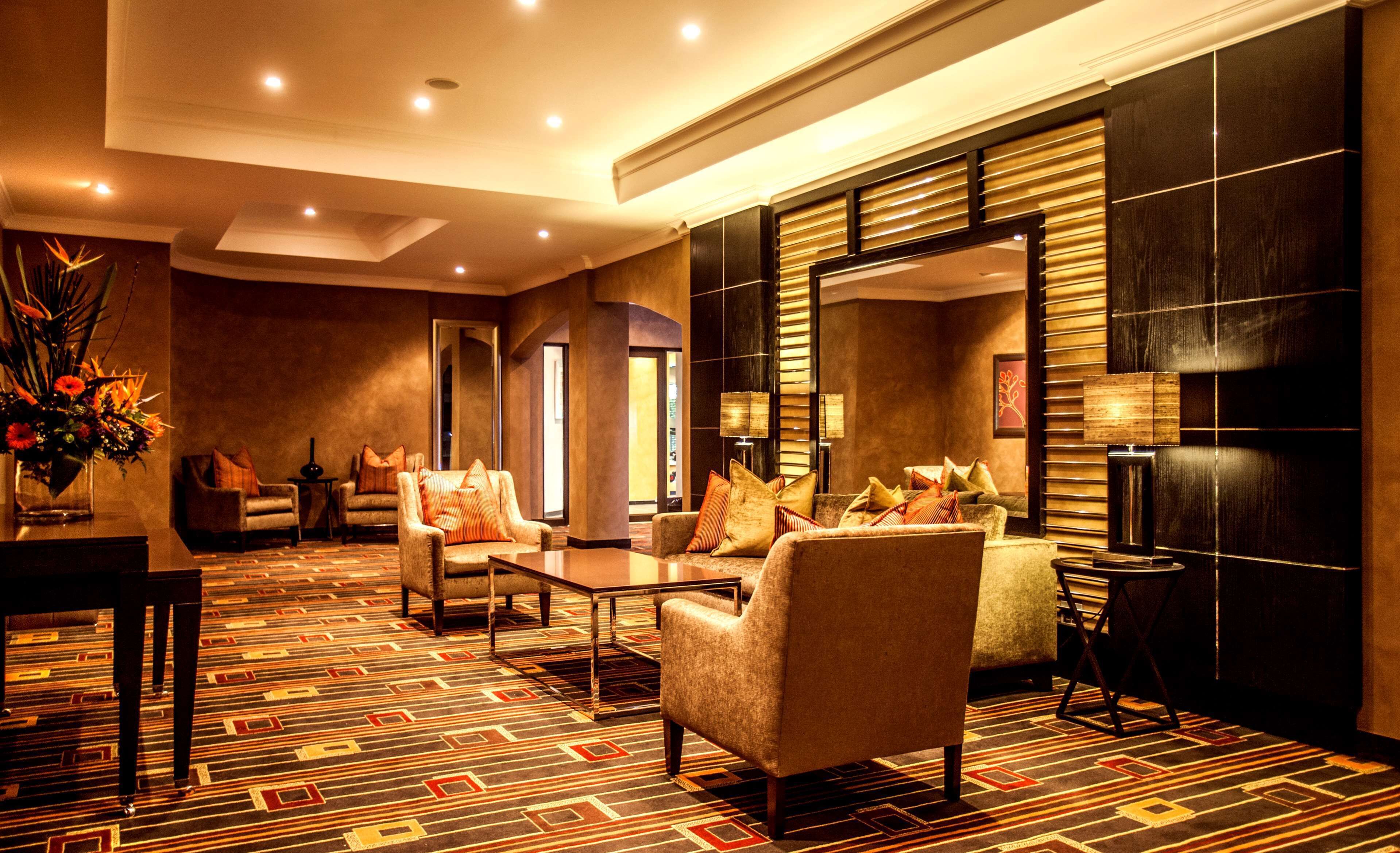 Un vestíbulo de hotel con alfombra estampada, área de estar con sillones y sofás, una mesa de centro, una mesita auxiliar con lámpara y un espejo grande.