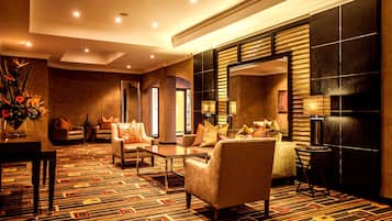 Un vestíbulo de hotel con alfombra estampada, área de estar con sillones y sofás, una mesa de centro, una mesita auxiliar con lámpara y un espejo grande.