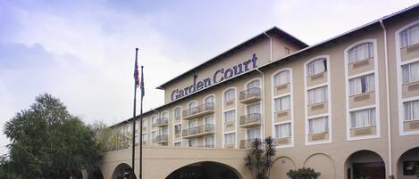 Ein Hotel namens Garden Court mit einer beigefarbenen Fassade, mehreren Balkonen und einem begrünten Eingang mit rosa Blumen.