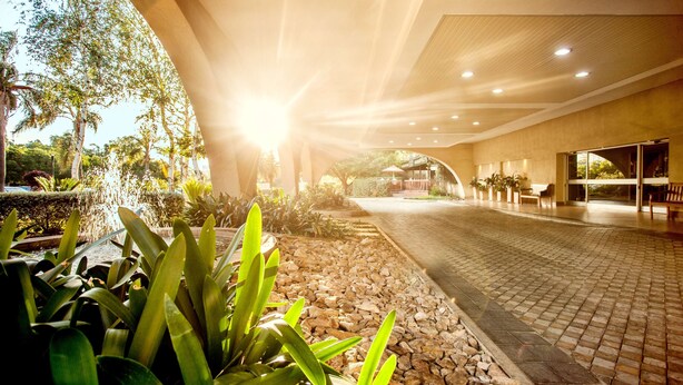 An indoor pathway with a fountain, surrounded by plants and a tiled floor, leading to a covered area with seating. - Garden Court O.R. Tambo International Airport (Kempton Park)