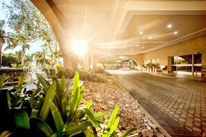 Un sendero interior con una fuente, rodeado de plantas y un piso de baldosas, que lleva a una zona cubierta con asientos.