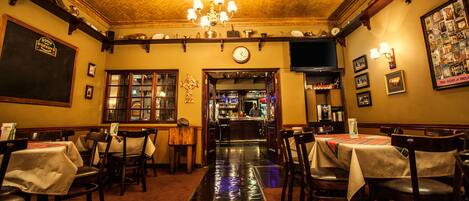 A restaurant with a bar area, wooden chairs, tables with white tablecloths, a chandelier, and a clock on the wall.