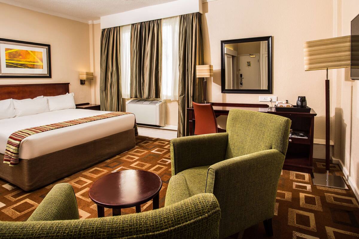 Quarto de hotel com uma cama grande, duas poltronas, uma mesa pequena, uma escrivaninha com abajur, um espelho e uma janela com cortinas.