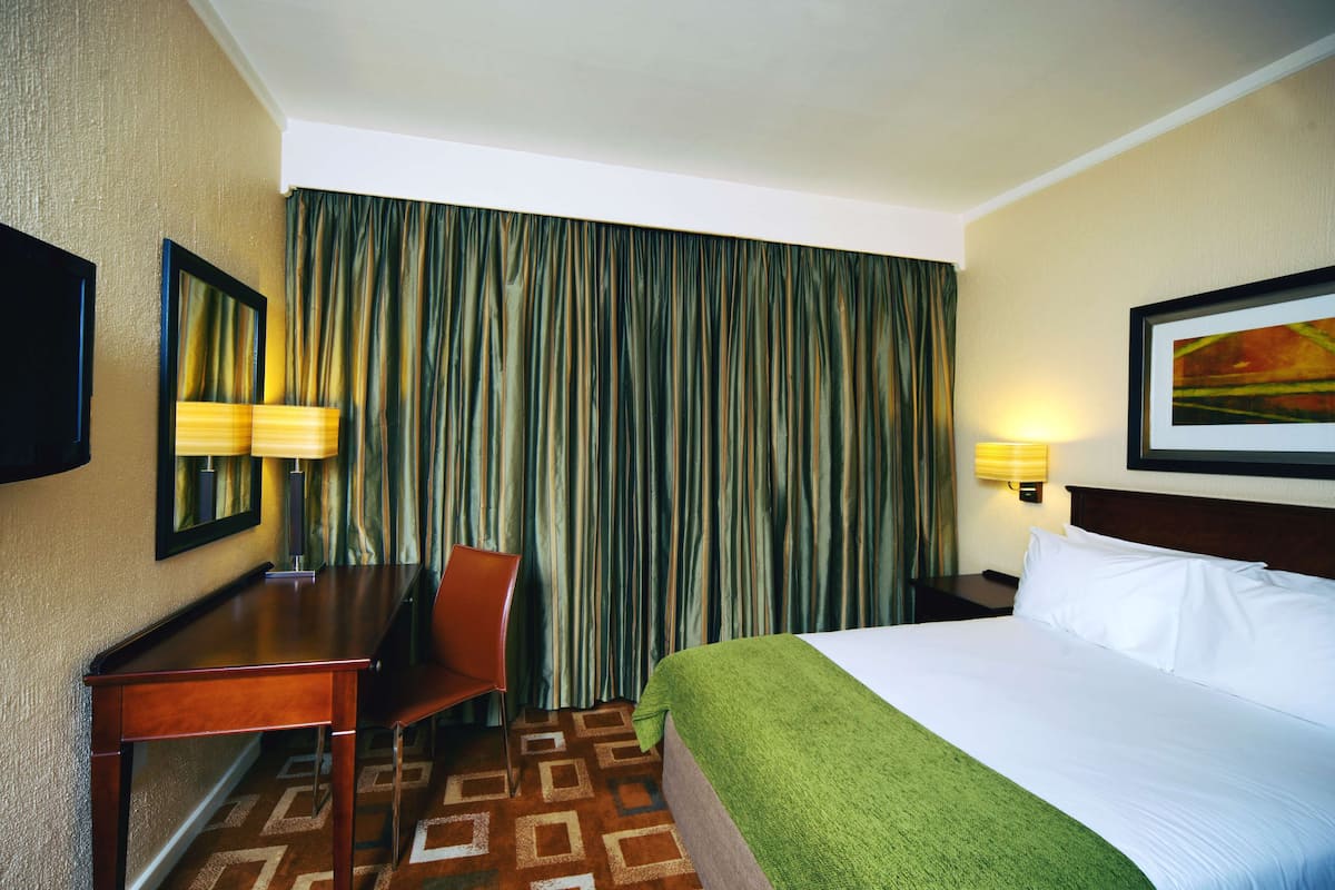 Quarto de hotel com uma cama, uma escrivaninha, uma cadeira, um abajur e uma janela grande com cortinas.