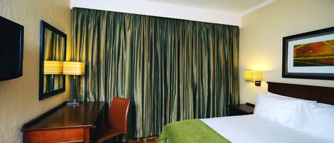 A hotel room with a bed, a desk, a chair, a lamp, and a large window with curtains.