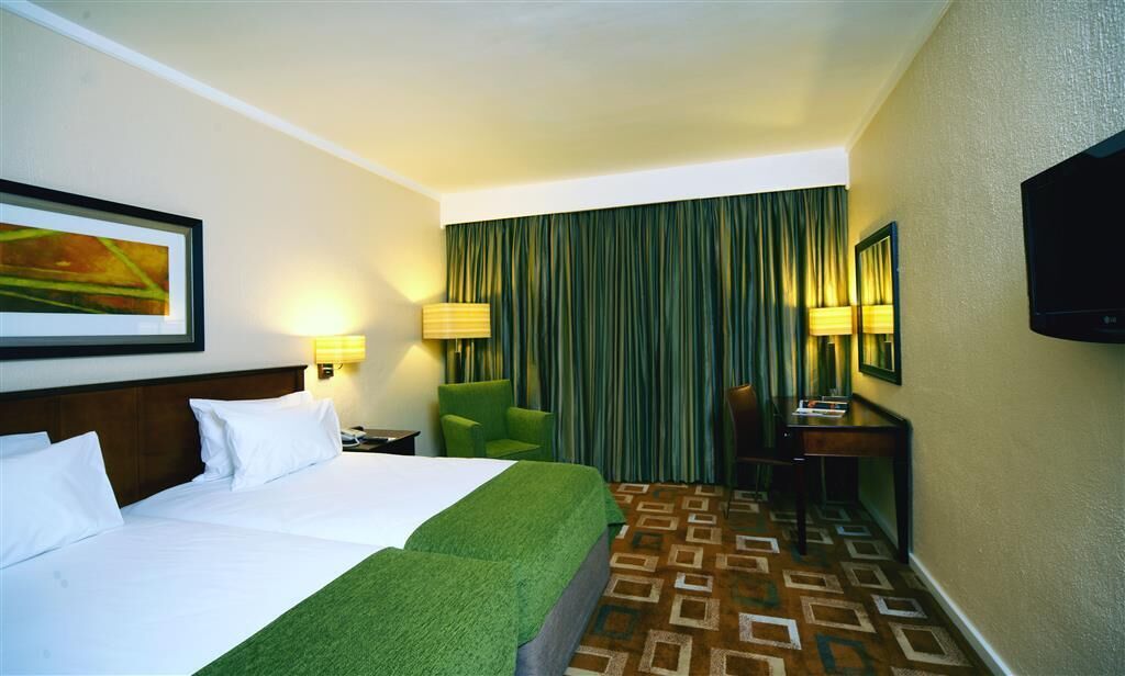 Habitación de hotel con dos camas, una silla verde, un escritorio, un televisor y una alfombra estampada.
