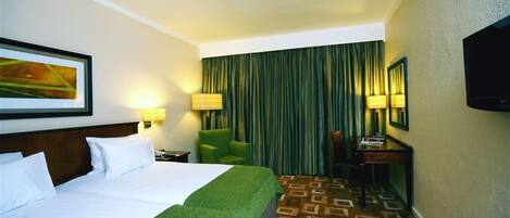 Habitación de hotel con dos camas, una silla verde, un escritorio, un televisor y una alfombra estampada.