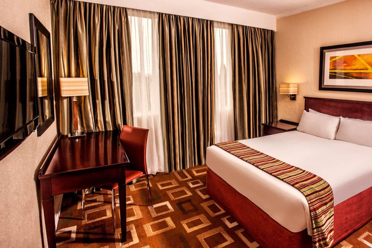 Quarto de hotel com uma cama grande, uma escrivaninha, uma cadeira, um telefone e um quadro na parede.