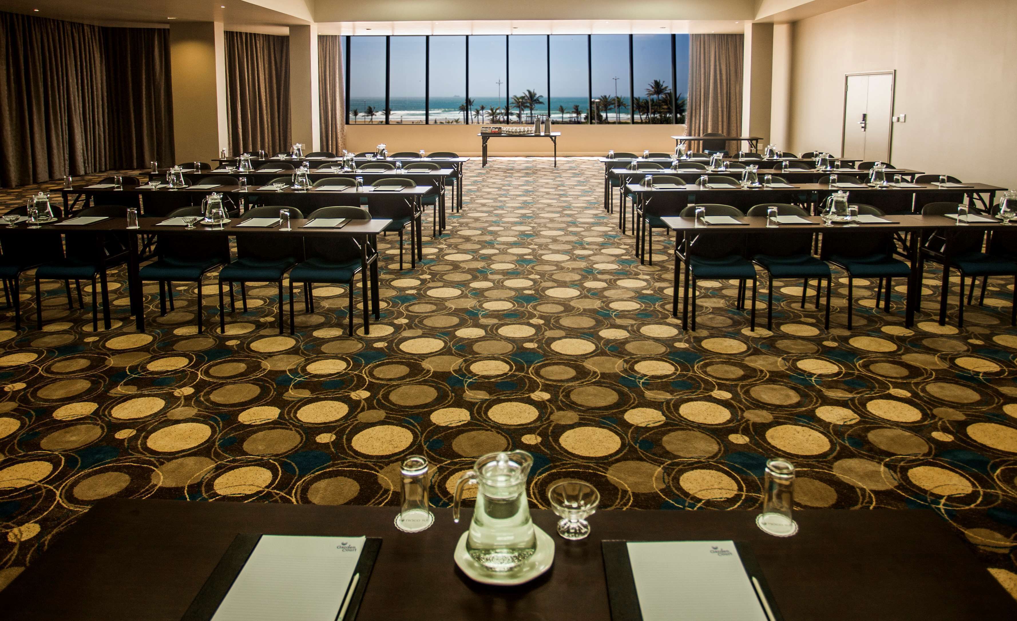 A conference room with a view of the beach, arranged tables with chairs, and a patterned carpet.