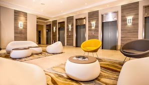 A modern lobby with a carpeted floor, several seating areas including a yellow chair, and a row of elevators.