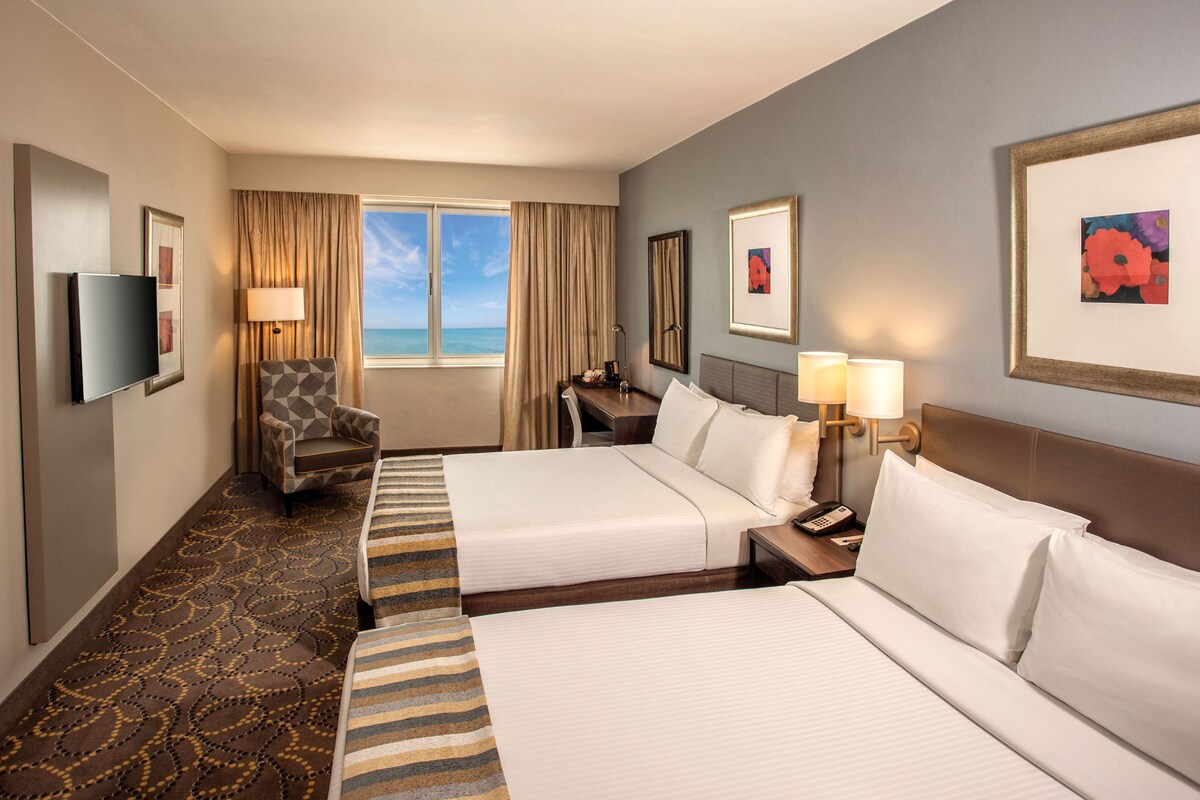 A hotel room with two beds, a desk, a chair, a TV, and a window with curtains.
