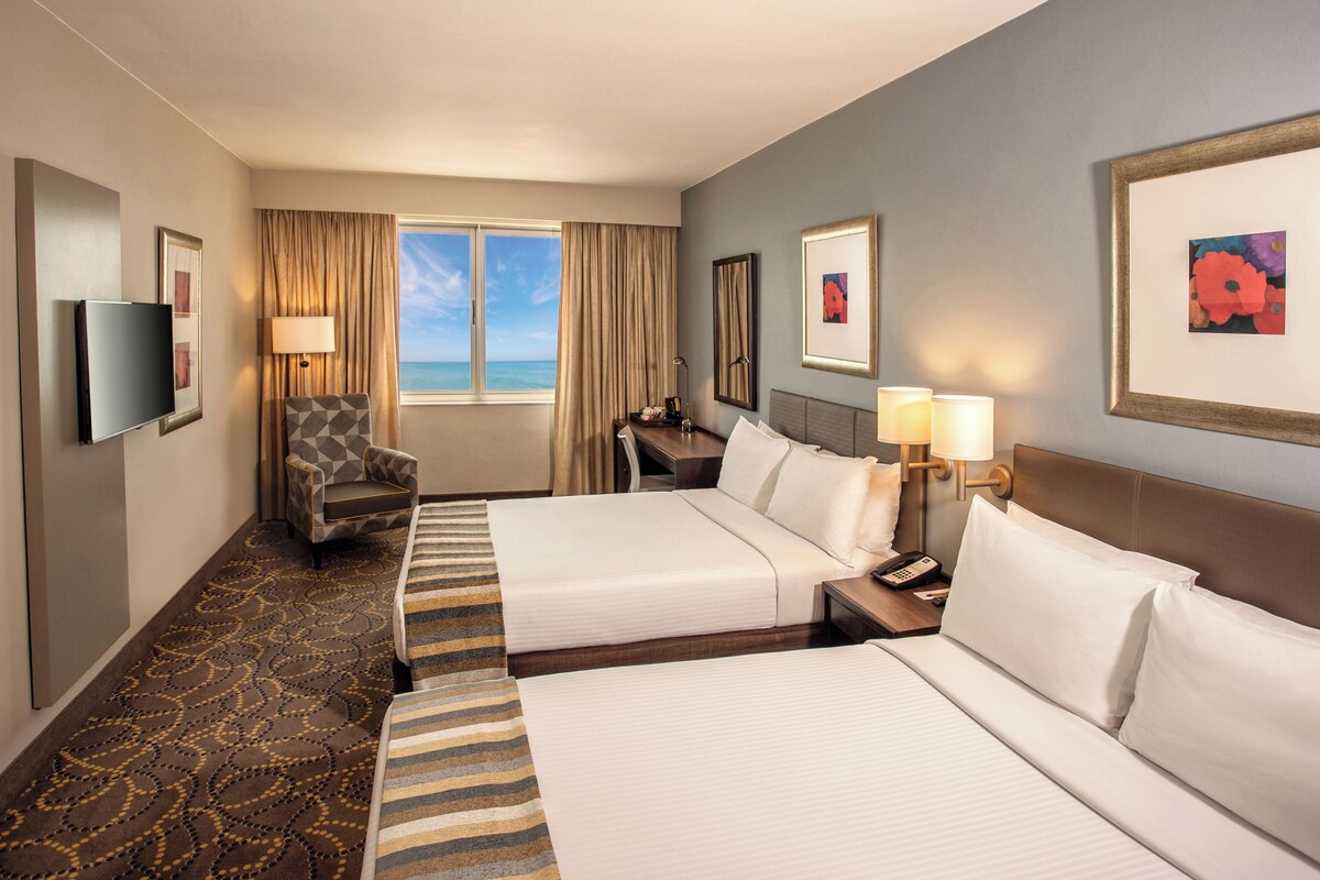 A hotel room with two beds, a desk, a chair, a TV, and a window with curtains.