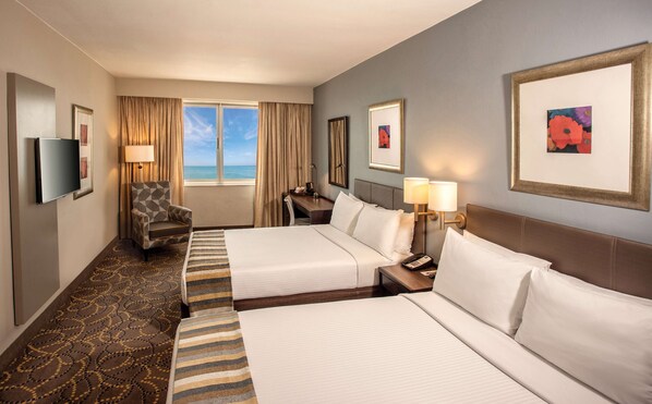 A hotel room with two beds, a desk, a chair, a TV, and a window with a view of the ocean. - Garden Court South Beach (Durban)