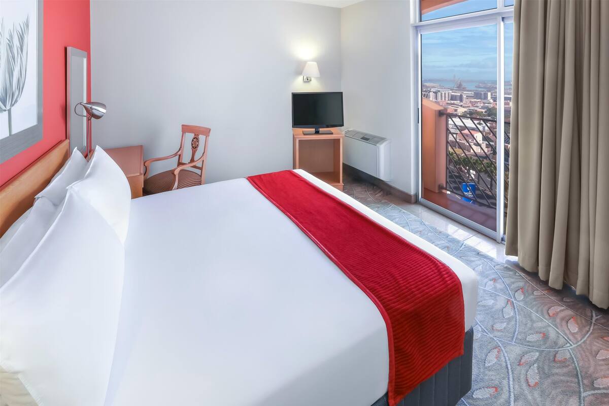 Une chambre d’hôtel avec un grand lit, un bureau avec une télévision et une fenêtre donnant sur la ville.