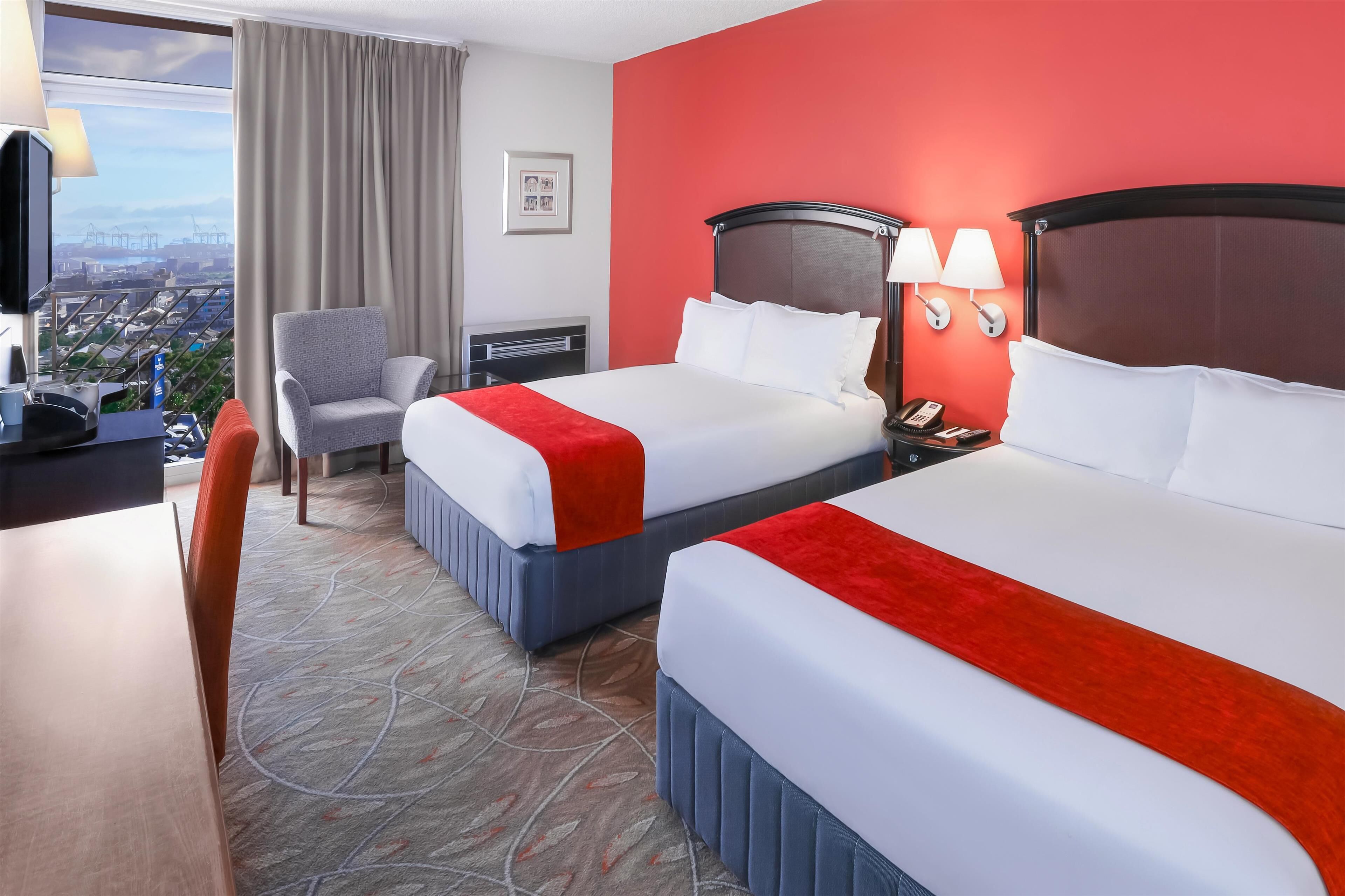 Een hotelkamer met twee bedden, een bureau, een stoel, een televisie en uitzicht op de stad.