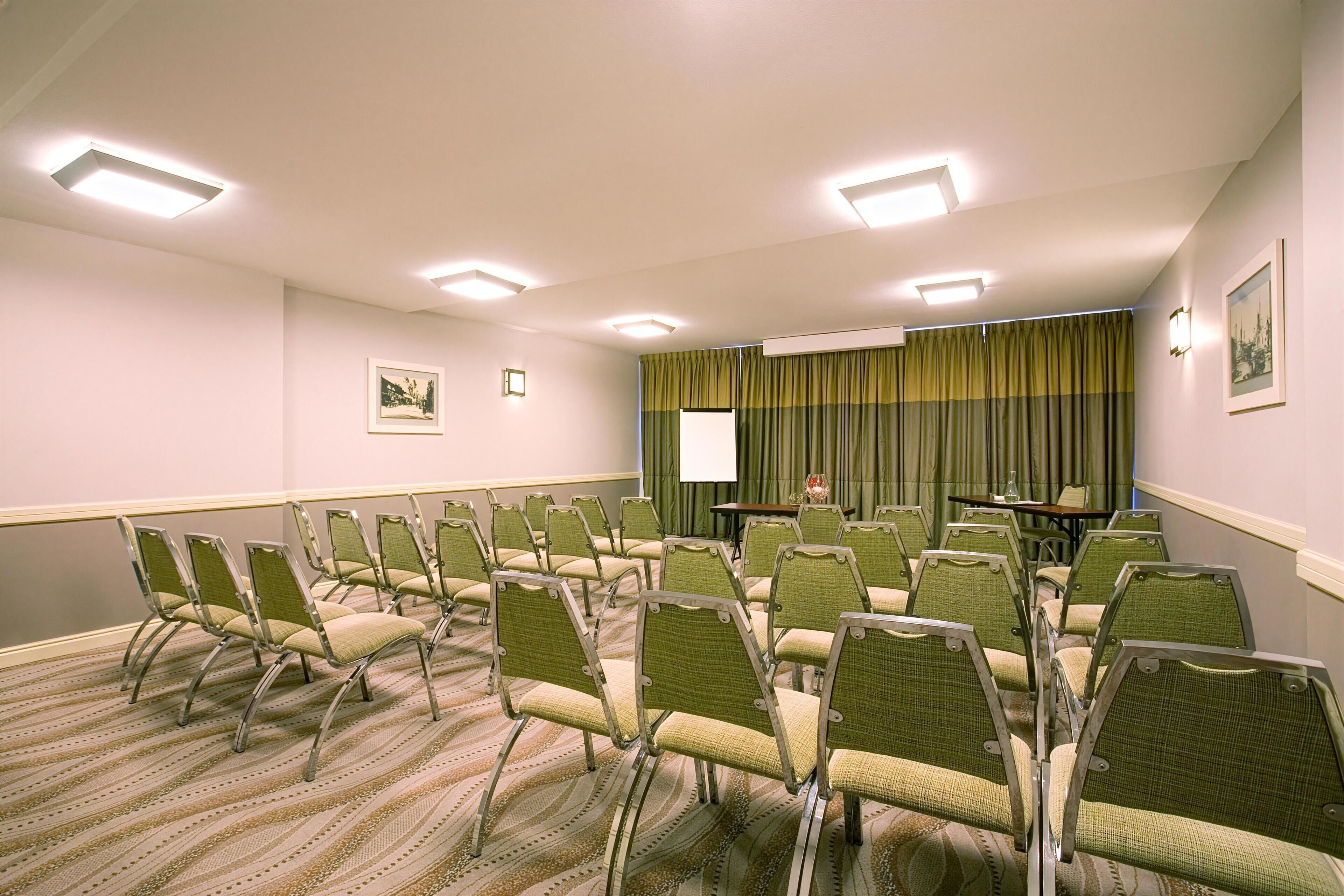 Une salle de conférence avec des rangées de chaises vertes, un écran de projection et une table sur laquelle est posé un vase.