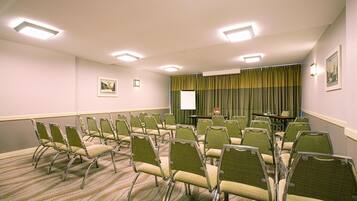 Une salle de conférence avec des rangées de chaises vertes, un écran de projection et une table sur laquelle est posé un vase.