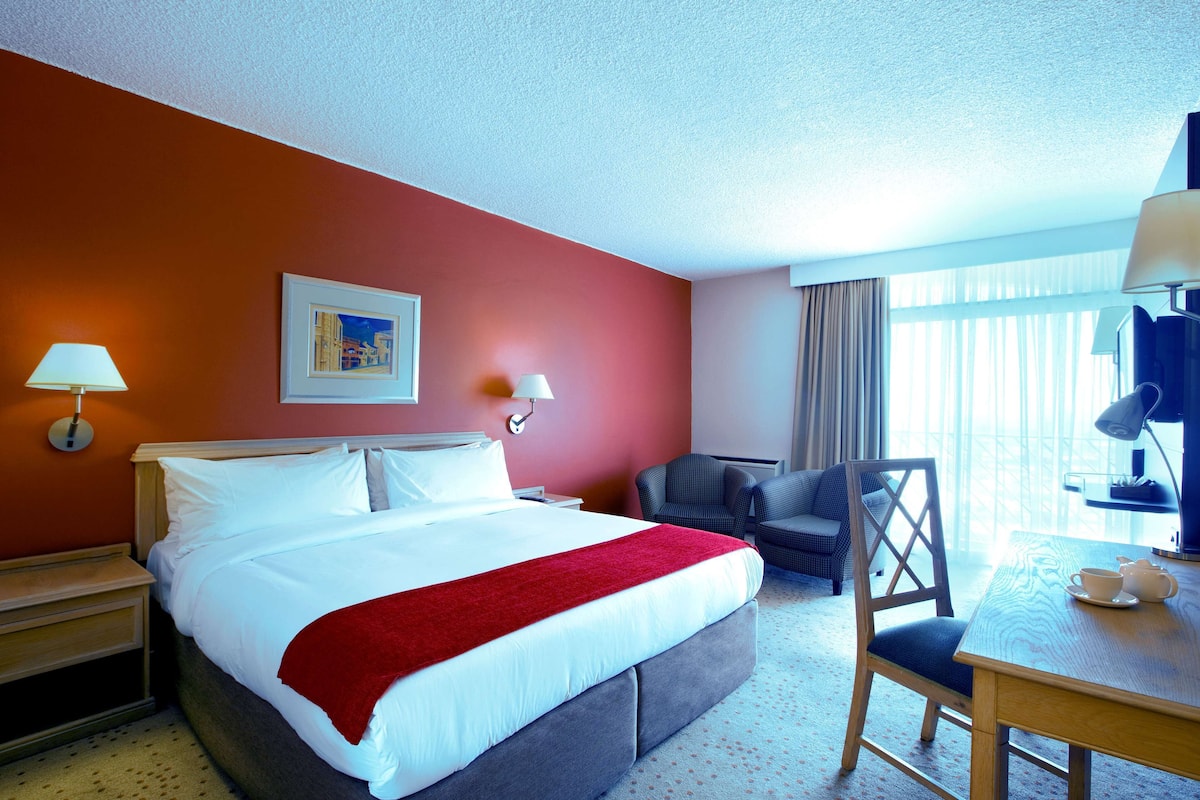 Une chambre d’hôtel avec un grand lit, un bureau avec une lampe, une chaise et une fenêtre avec des rideaux.