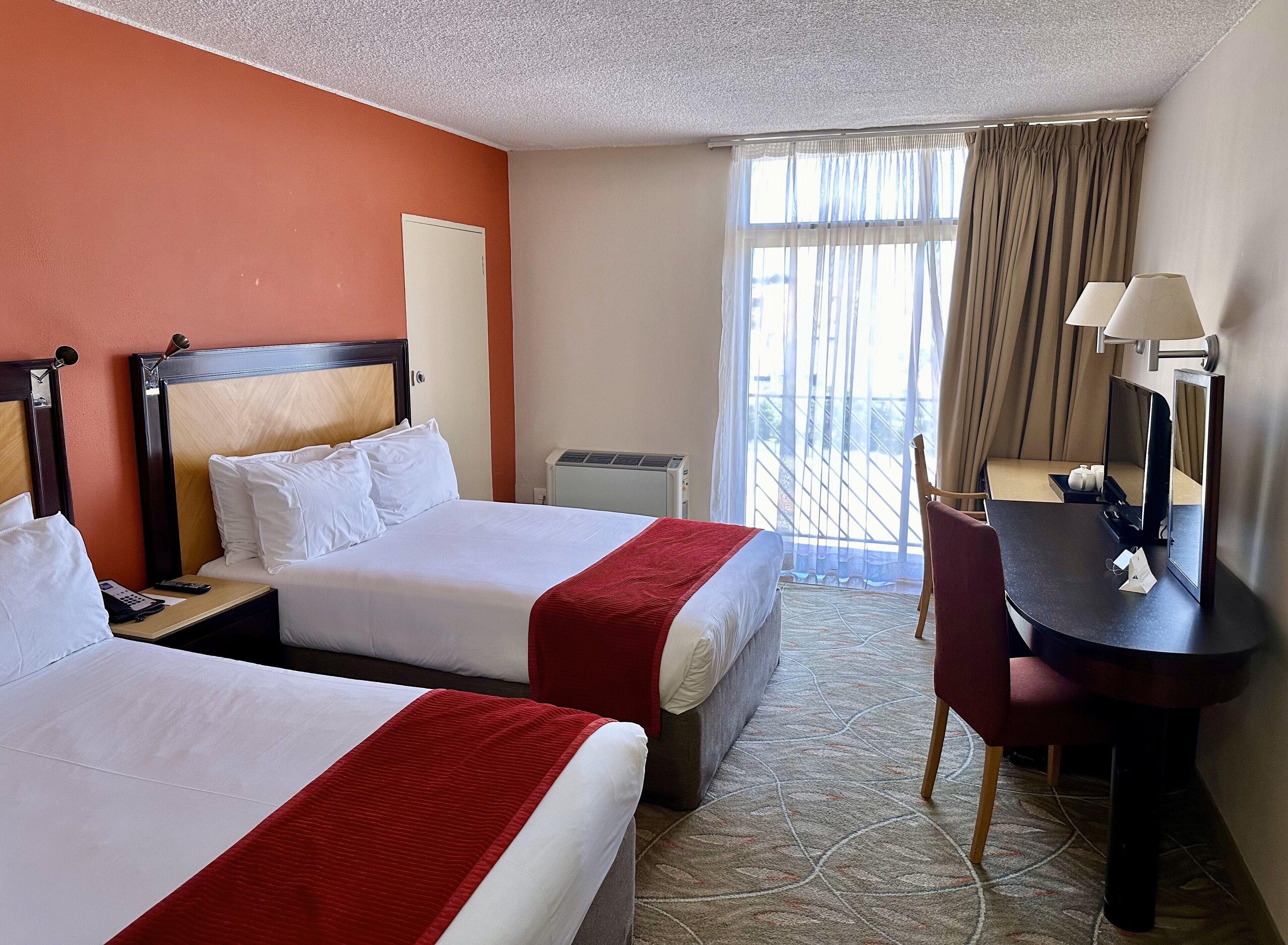A hotel room with two beds, a desk with a chair, a window with curtains, and a wall-mounted lamp.