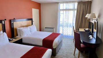 A hotel room with two beds, a desk with a chair, a window with curtains, and a wall-mounted lamp.