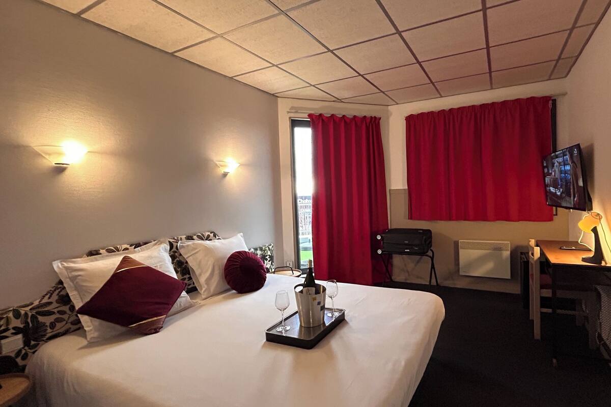 A hotel room with a bed, a desk, a TV, and a window with red curtains.