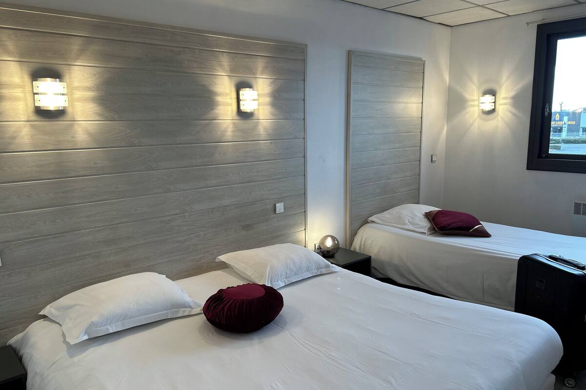 A hotel room with two beds, each with white linens and a red accent pillow. There are wall-mounted light fixtures and a window with a view of the outside.