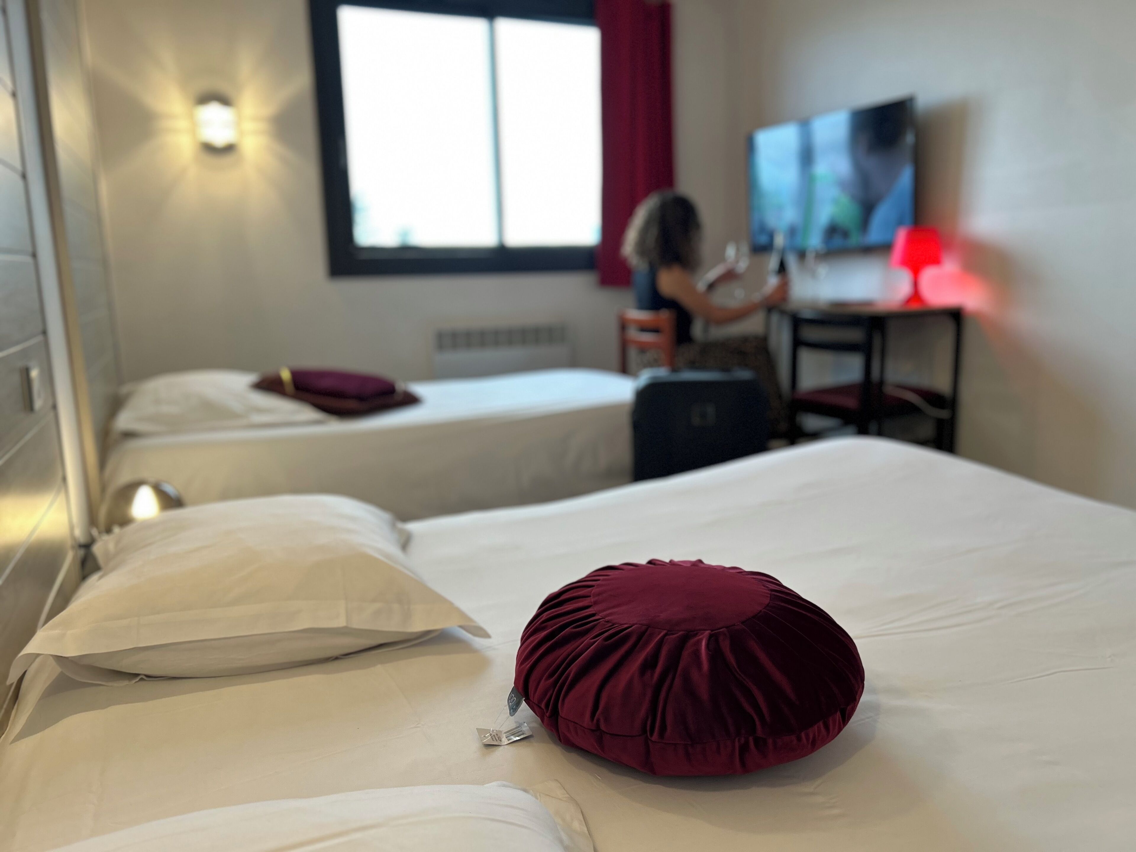 Habitación de hotel con dos camas individuales, un escritorio con silla, televisor y ventana con cortinas.