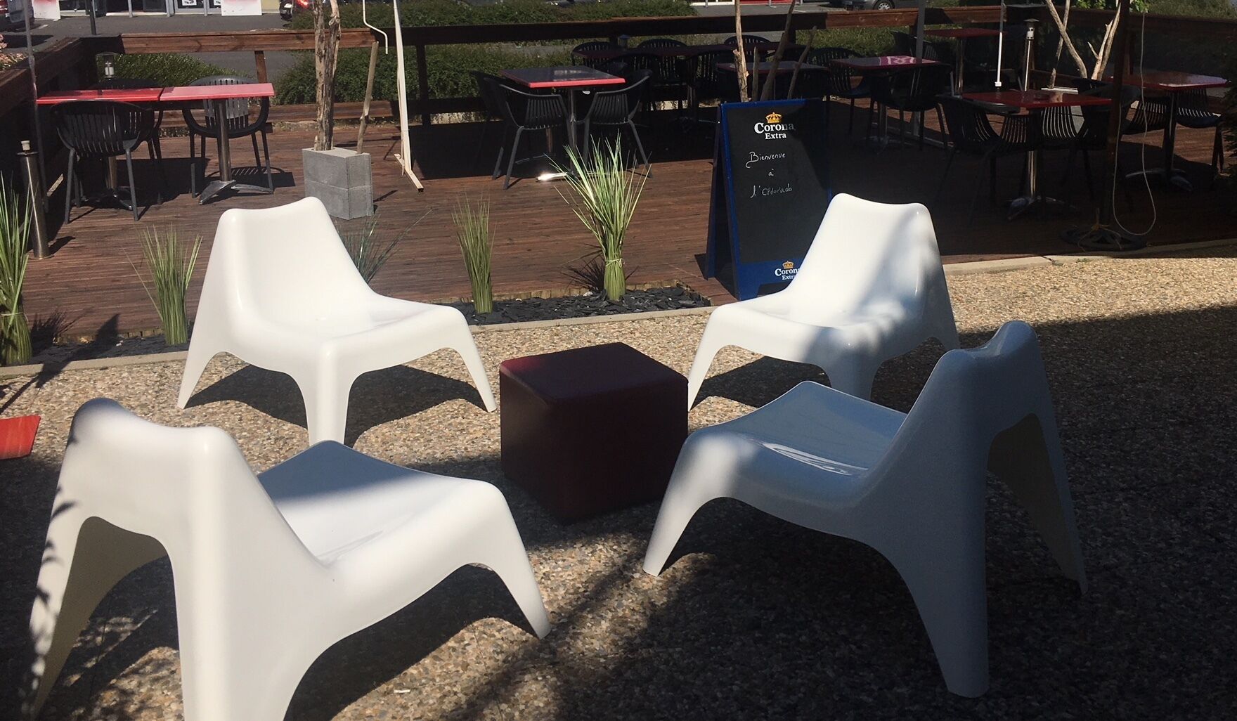 Sitzgelegenheit im Freien mit weißen Plastikstühlen und einem roten Tisch, umgeben von einer Holzterrasse und Kies.