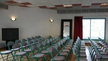 Un salón de conferencias con filas de sillas plegables, un televisor de pantalla plana grande y dos lámparas fijadas a la pared.