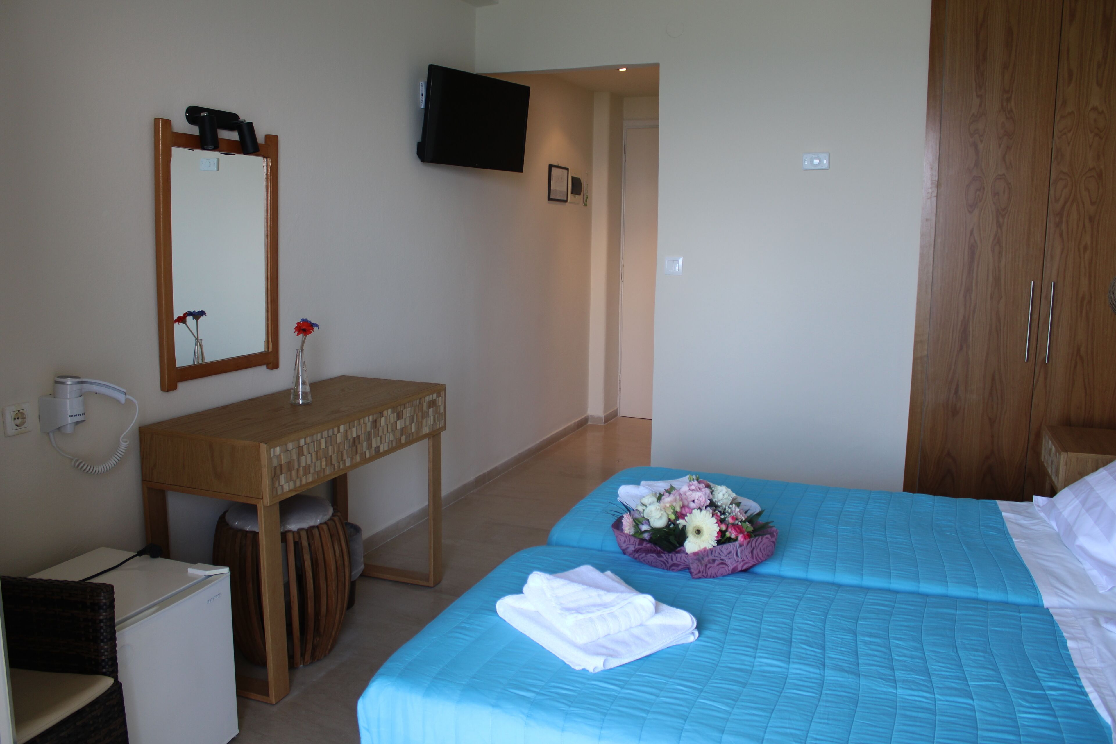 A hotel room with a wooden desk, a mirror, a television mounted on the wall, a bed with blue bedding, and a small trash bin.
