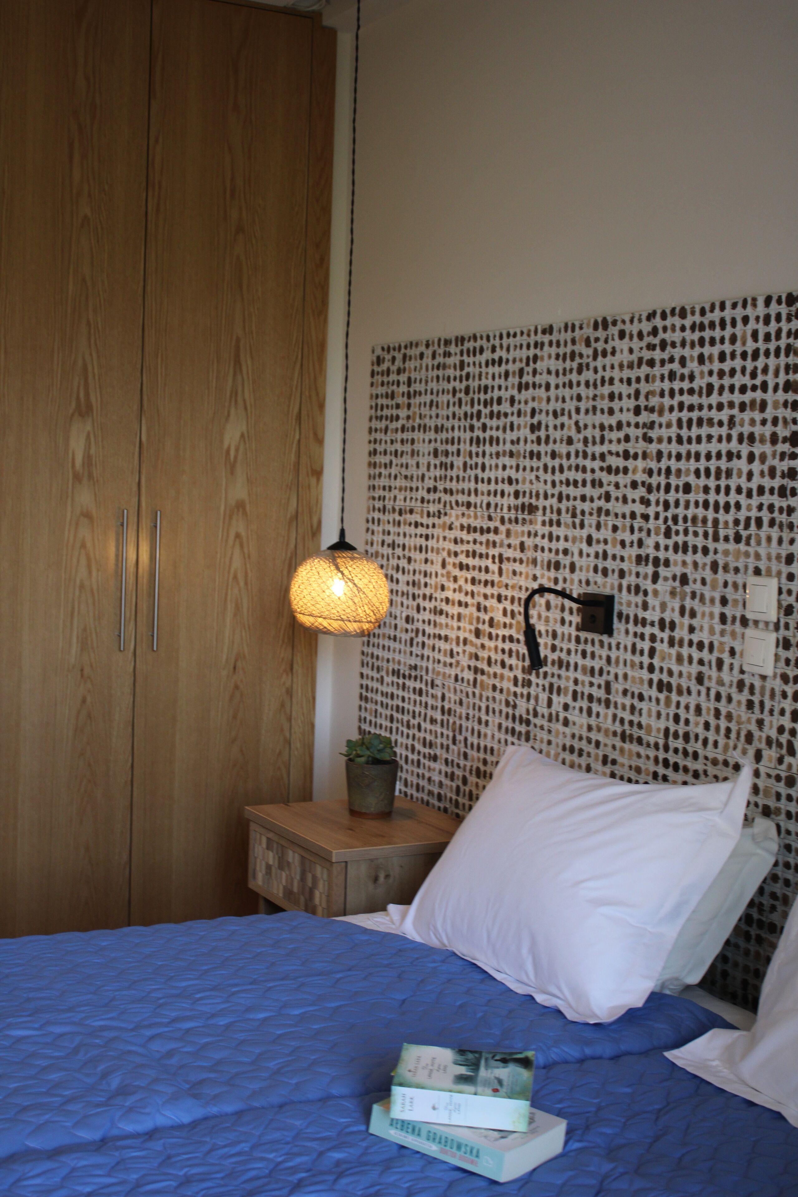 A bedroom with a wooden wardrobe, a bedside table with a lamp, a potted plant, and a bed with a blue bedspread and white pillows.