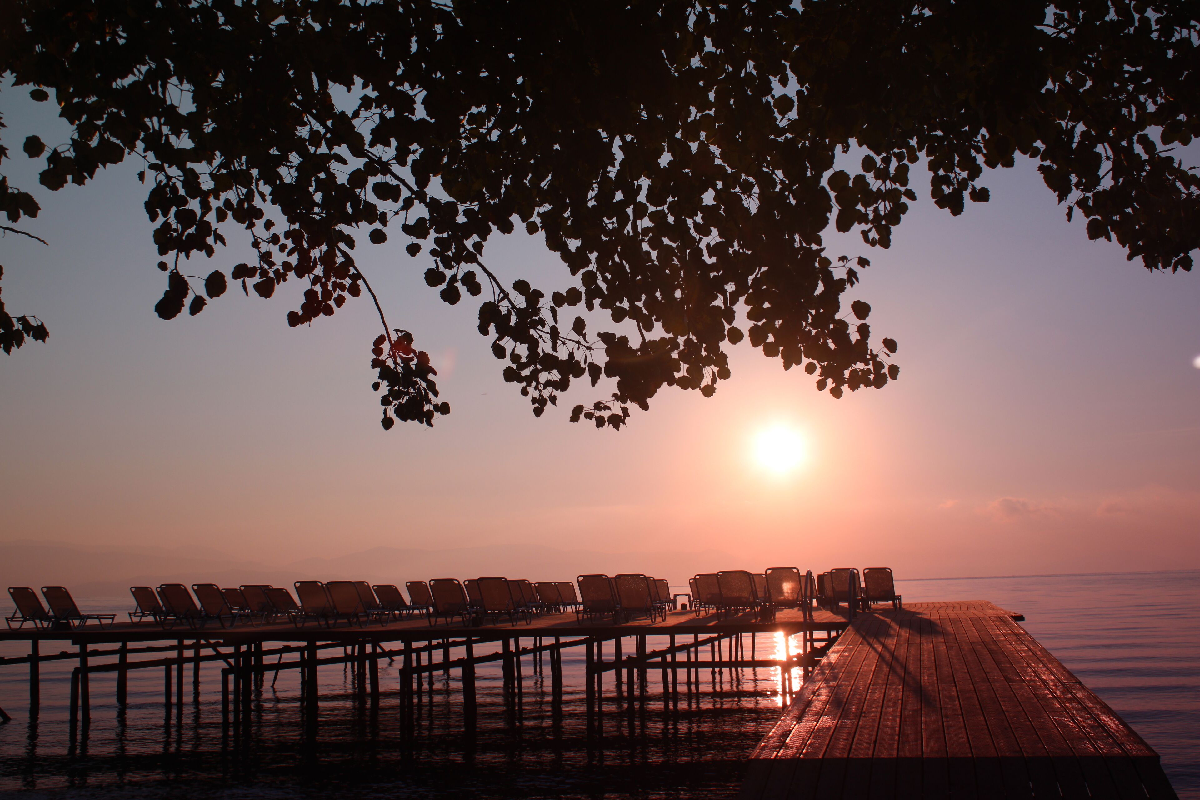 A wooden pier with rows of empty lounge chairs facing a sunset over calm water.