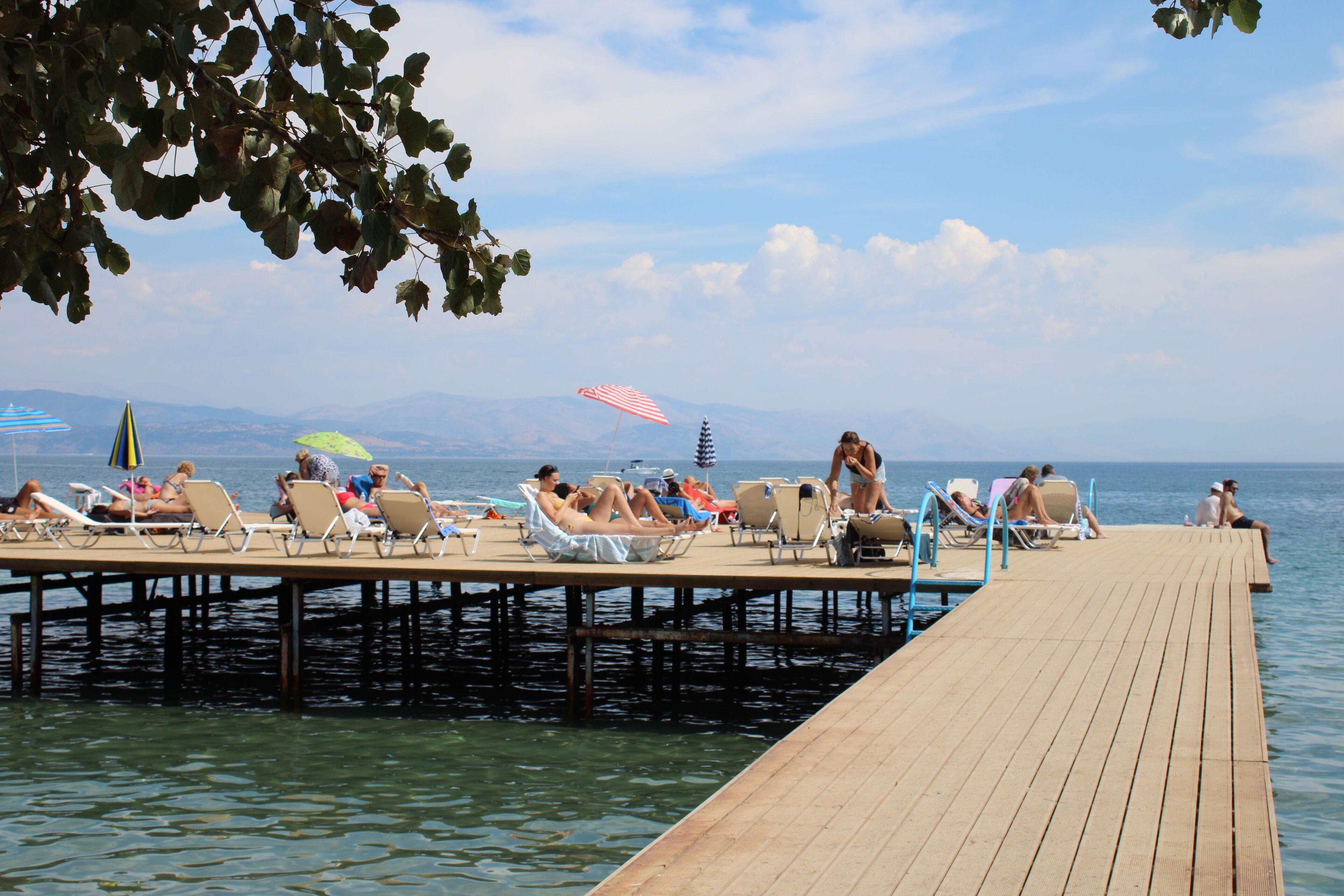 Een houten steiger met ligbedden en parasols, die zich uitstrekt over kalm water met bergen op de achtergrond.