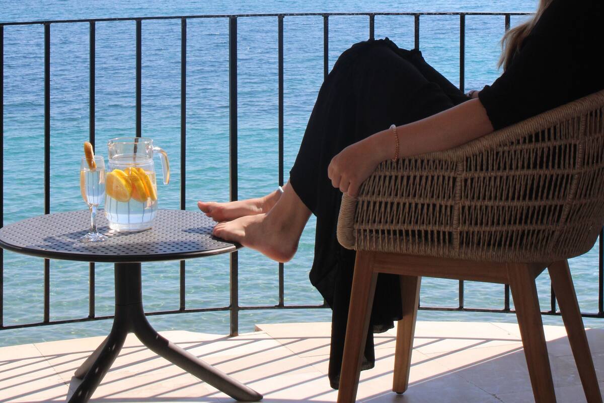A person seated on a wicker chair overlooking a body of water, with a table in front of them holding a drink and a glass.