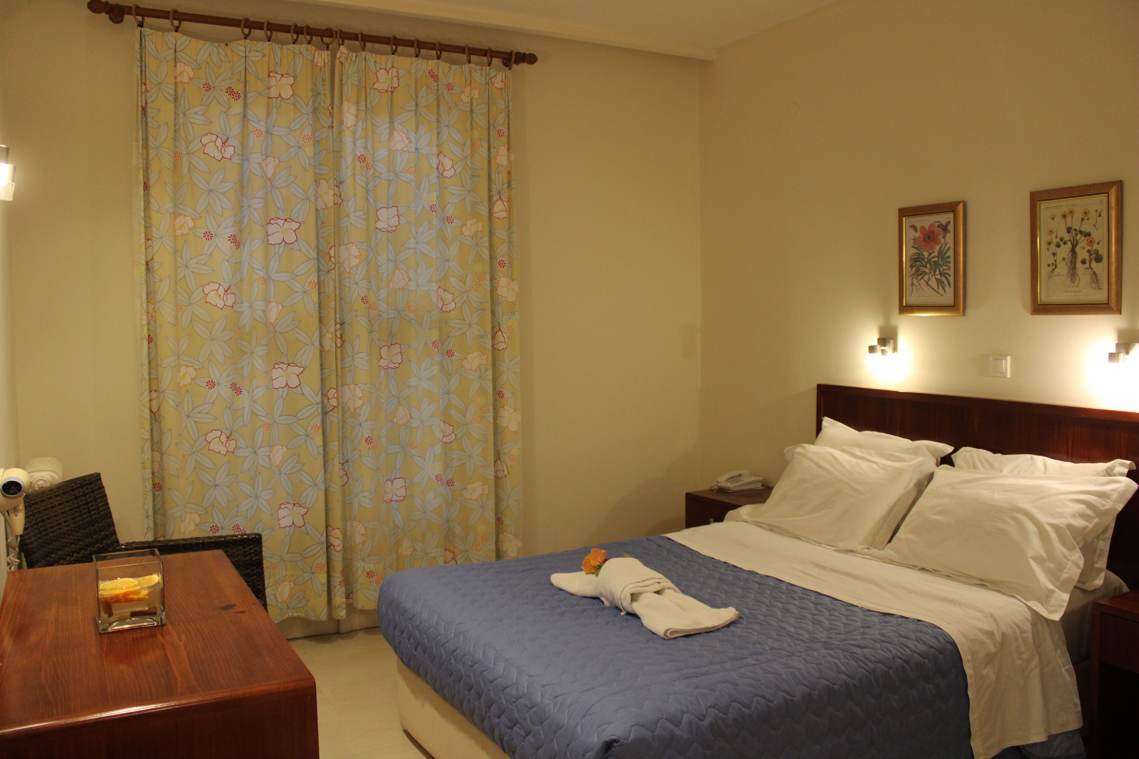 A hotel room with a bed, a wooden table, a chair, a desk, a telephone, and a curtain with a floral pattern.