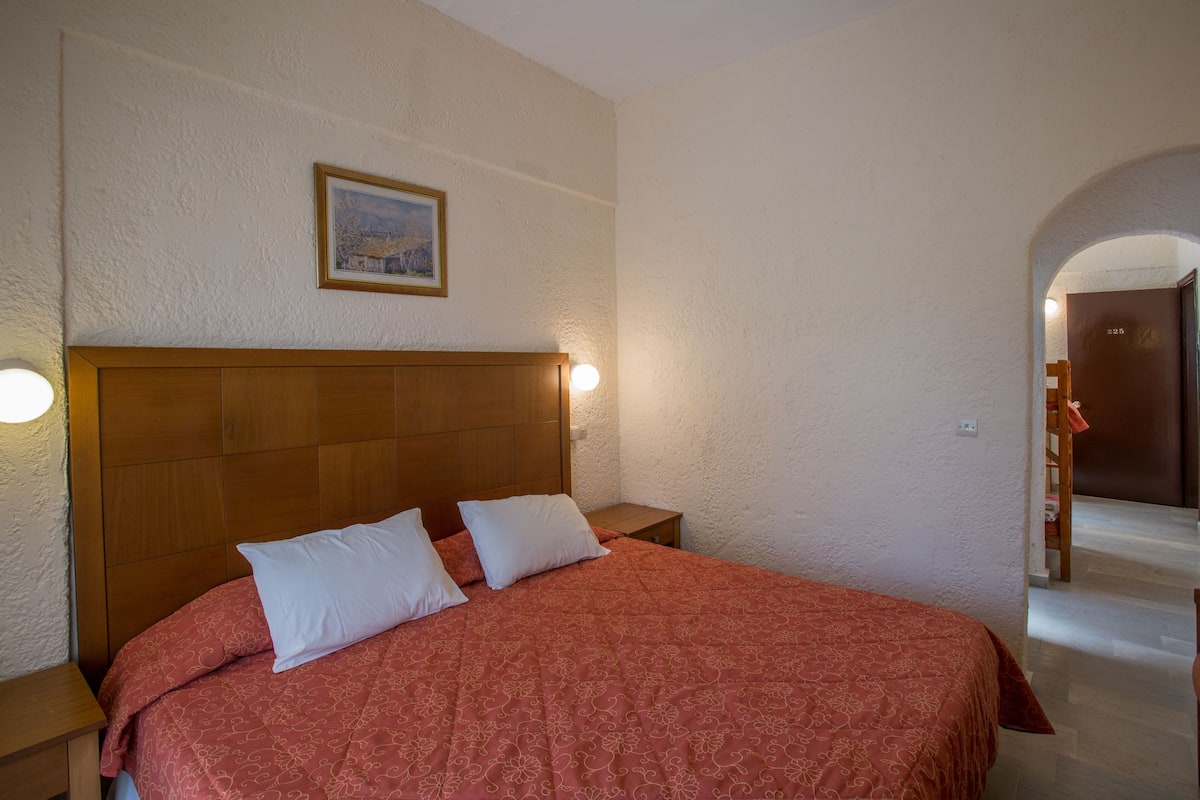 Un dormitorio con cabecera de madera, colcha estampada roja, dos almohadas blancas, una mesita de noche con lámpara y un cuadro en la pared.
