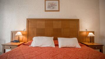 Una cama bien hecha con una colcha roja estampada, dos almohadas blancas y un cabecero de madera. Hay dos mesitas de noche con lámparas a cada lado de la cama.