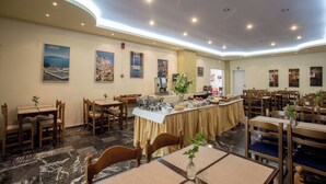Uma área de jantar de hotel com piso de mármore, mesas e cadeiras de madeira, um serviço de buffet e obras de arte nas paredes retratando a Grécia.