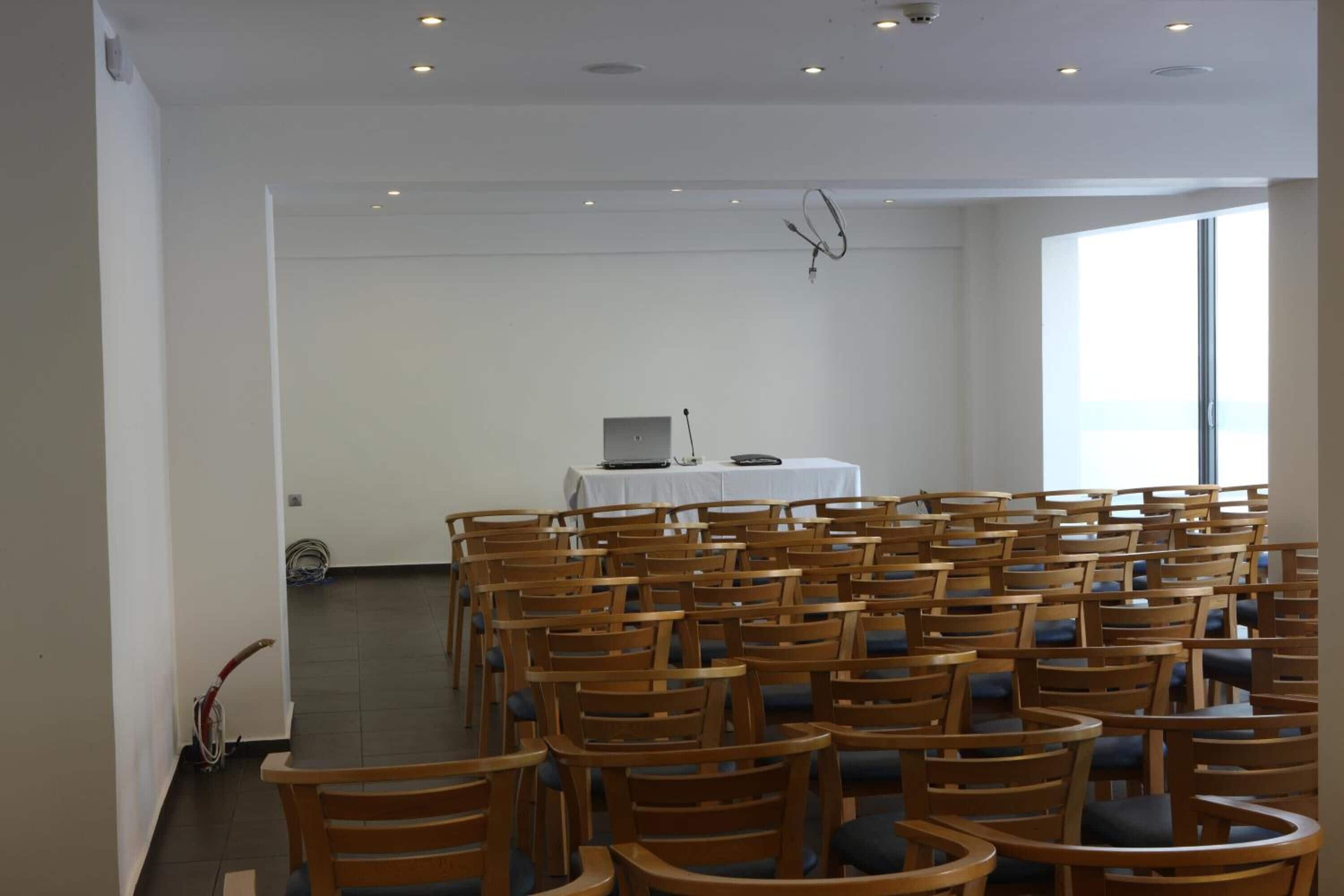 A conference room with rows of wooden chairs, a white table with a laptop, and a large window providing natural light.