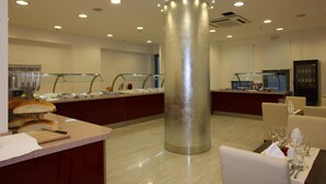 Un espace de restauration moderne dans un hôtel, avec un pilier central, des comptoirs de buffet et un coin salon avec des tables dressées pour le repas.