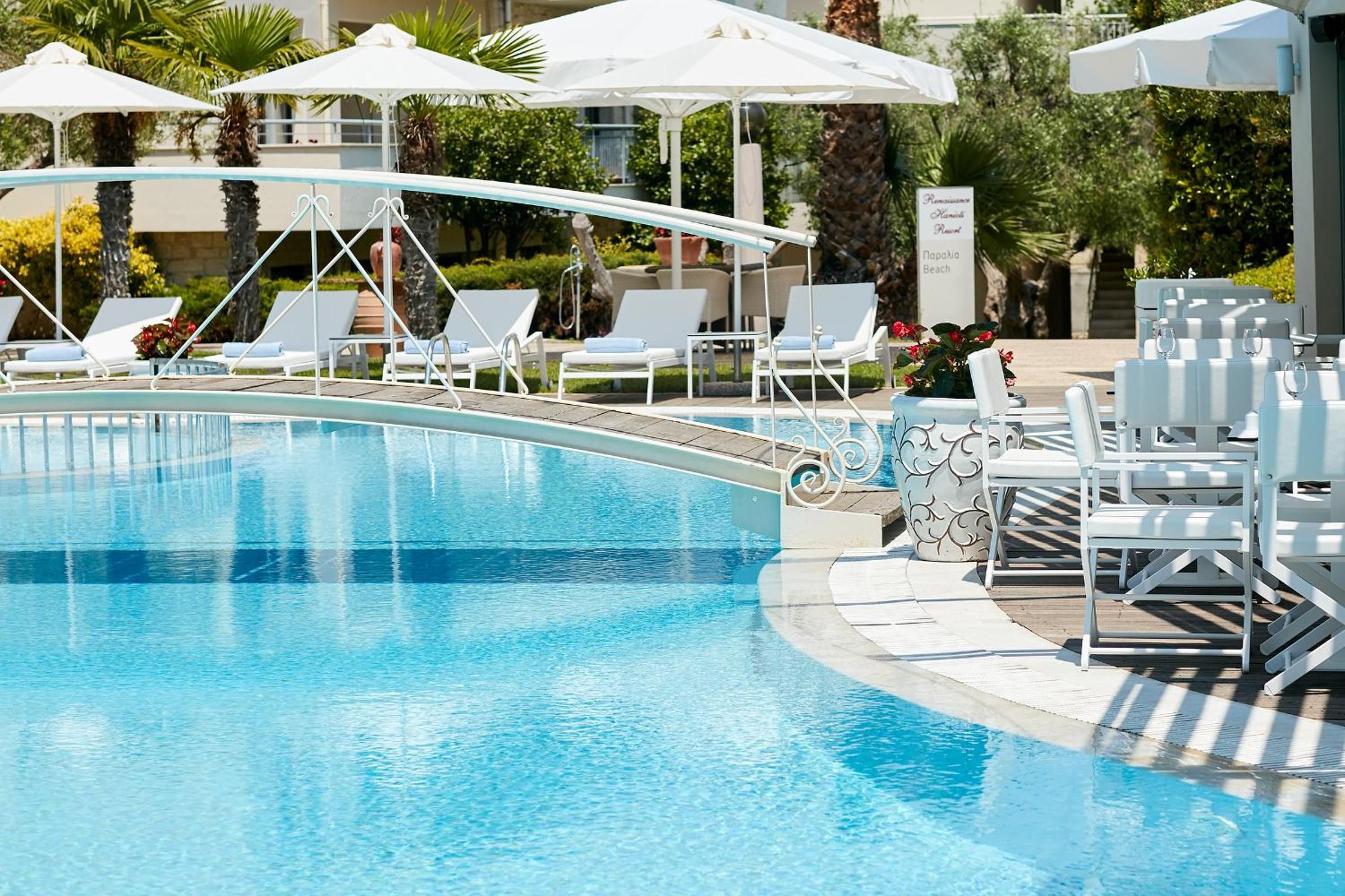 A swimming pool with lounge chairs and umbrellas, surrounded by greenery and a building.