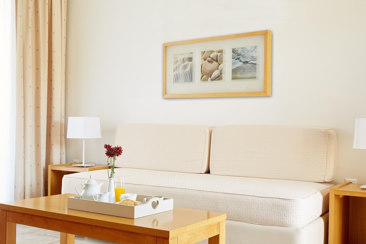 A hotel room with a wooden table, a white sofa, a framed picture on the wall, and a window with curtains.