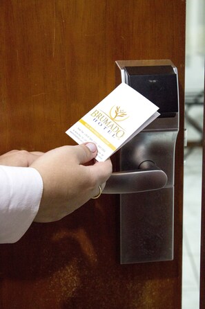A hand holding a business card in front of a door with a metallic handle. - Brumado Hotel (Campo Grande)