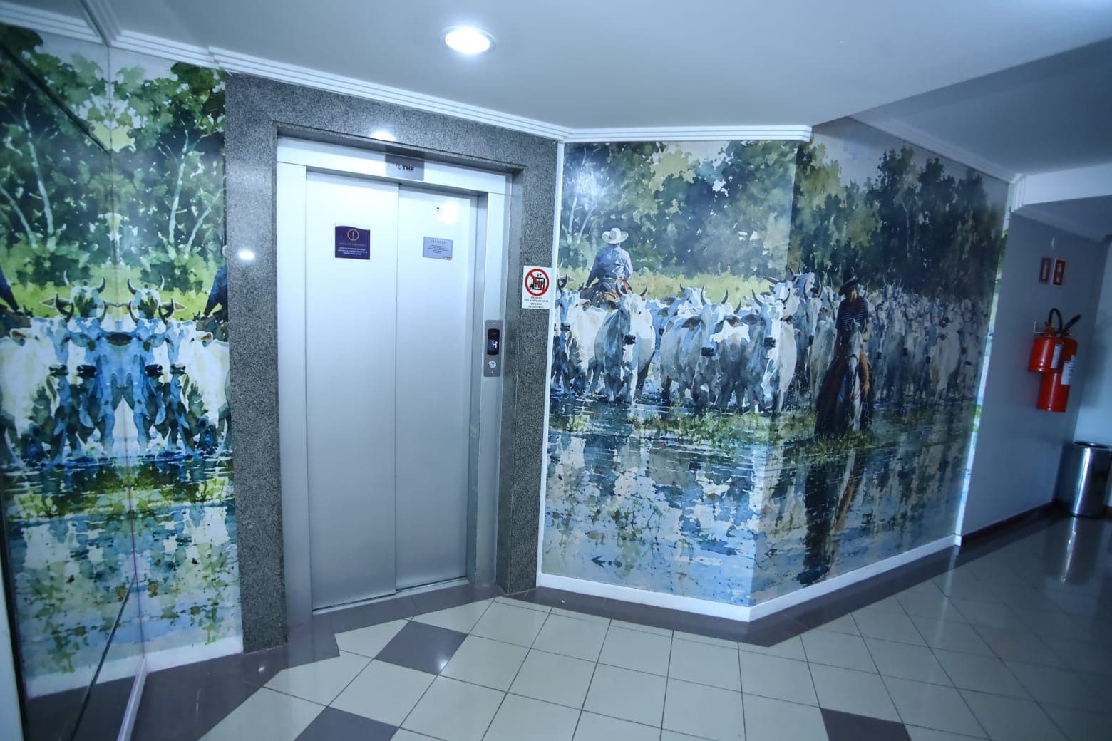 Un ascenseur orné d’un panneau « interdiction de fumer », d’un extincteur et d’une fresque murale représentant un troupeau de bovins.