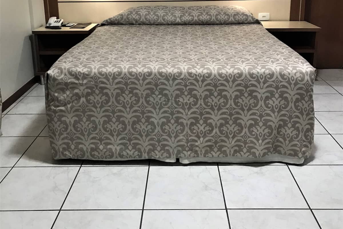 A hotel room with a bed, bedside tables, a wall-mounted air conditioner, a painting, and a door.