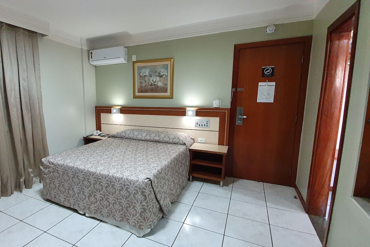 A hotel room with a bed, a nightstand, an air conditioning unit, a door, and a painting on the wall.