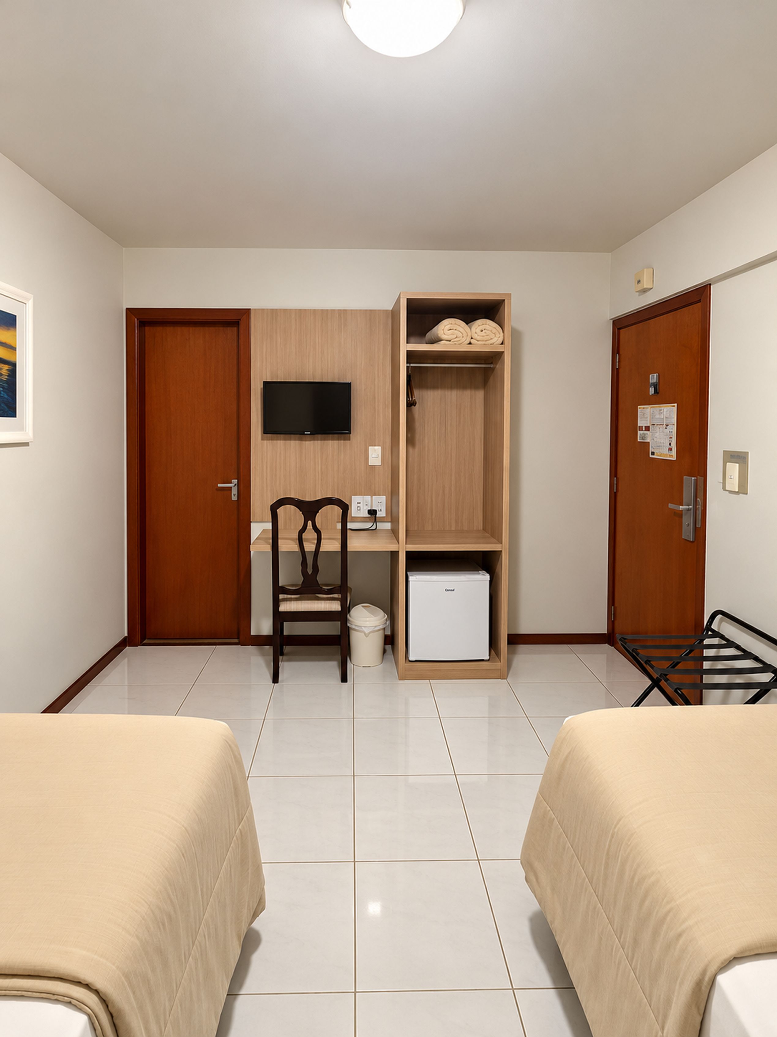 A hotel room with two beds, a desk, a chair, a television, a wardrobe, and a small refrigerator.
