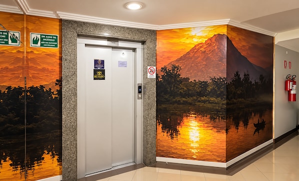 An elevator with a wall mural depicting a sunset over a mountain and a lake. - Brumado Hotel (Campo Grande)
