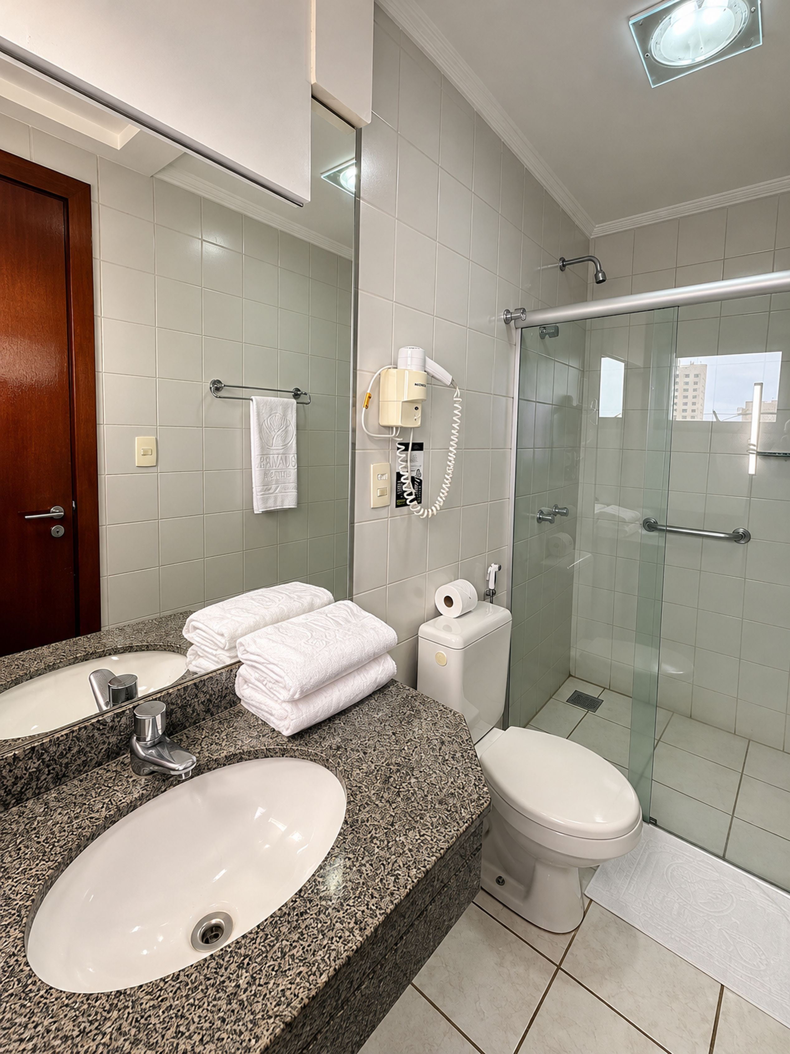 A bathroom with a granite countertop, two sinks, a toilet, a glass shower enclosure, and a towel rack with a white towel.